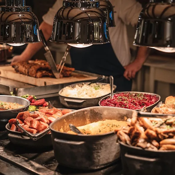 En person skjærer kjøtt ved en travel buffet med ulike varme retter og salater under varmelamper.