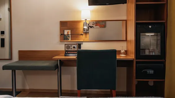 A cozy hotel room desk area with a chair, lamp, radio, mini-fridge, and safe, offering a comfortable stay at Home Hotel Kung Oscar.