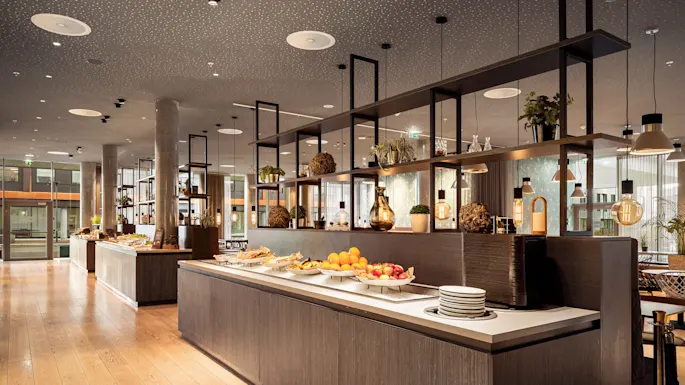 Buffet tables displaying fruits and various dishes are arranged in a modern dining area with wooden floors and decorative lighting.