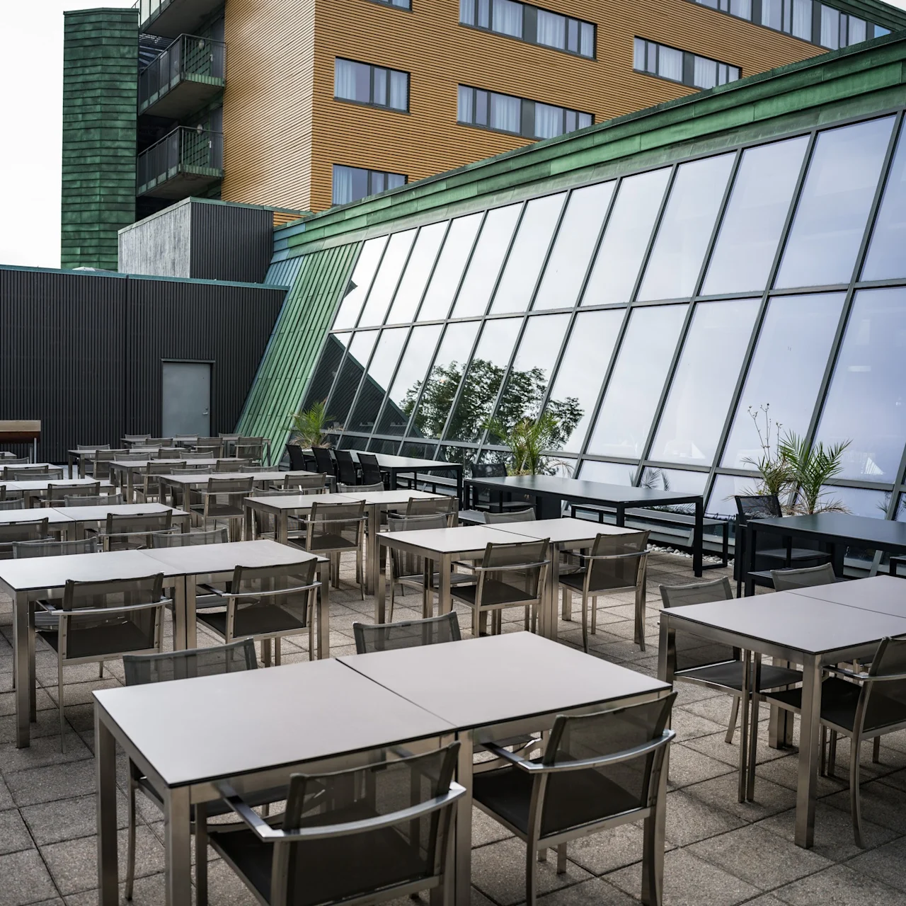 Bord och stolar är prydligt arrangerade på en takterrass, intill en modern byggnad med stora vinklade fönster, omgivna av några krukväxter.