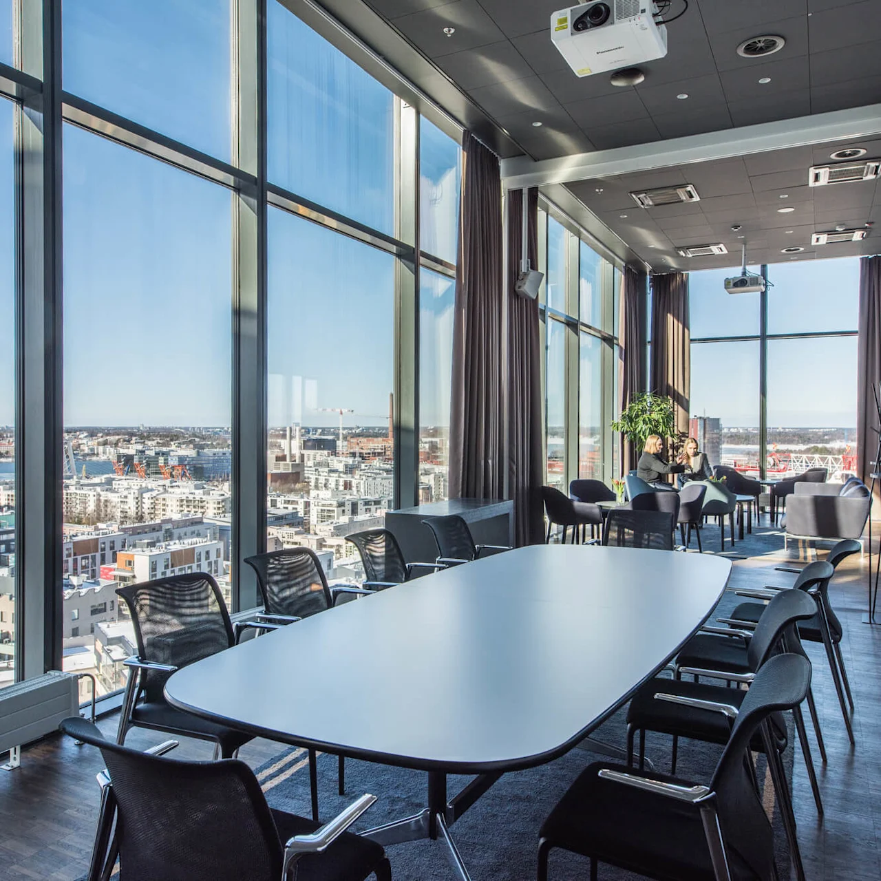 Et rummeligt konferencelokale har et langt bord omgivet af stole. Store vinduer tilbyder panoramaudsigt over byen. I nærheden sidder to personer og samtaler i en hyggelig siddegruppe.