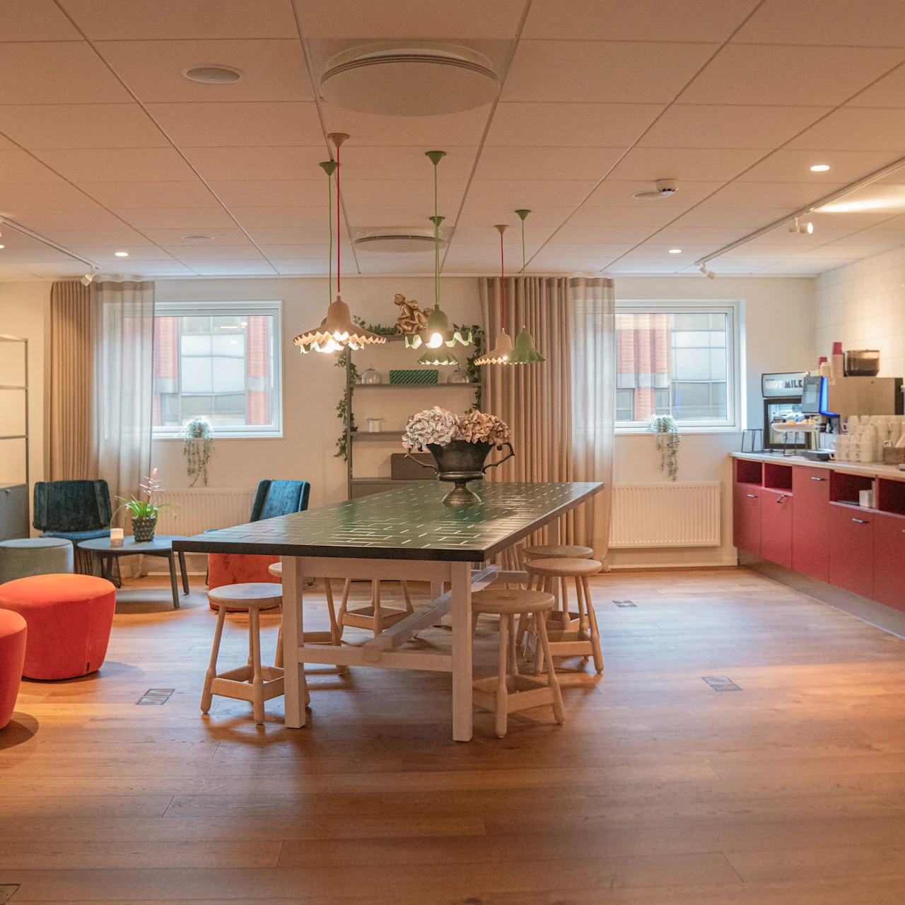 Et rummeligt køkken med et langt bord og taburetter i midten, omgivet af farvestrålende siddepladser og en væg med røde skabe. Lys belysning og store vinduer fremhæver den hyggelige atmosfære.