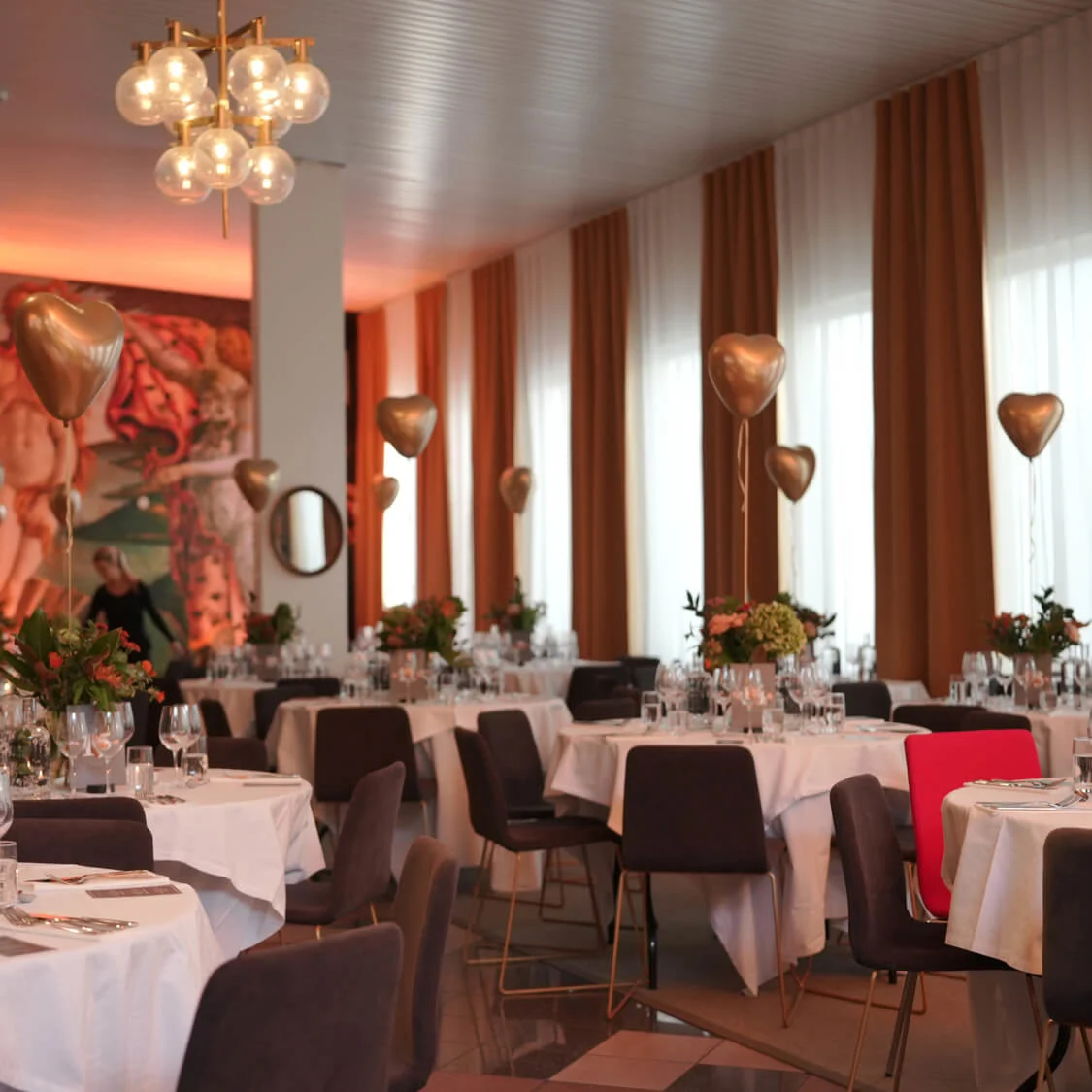 Luksuriøse spiseborde omsluttet af hvide duge har blomsterdekorationer og hjerteformede guldballoner. Elegante spisestole omgiver dem i et varmt oplyst rum, prydet med kunstværker og gardiner.