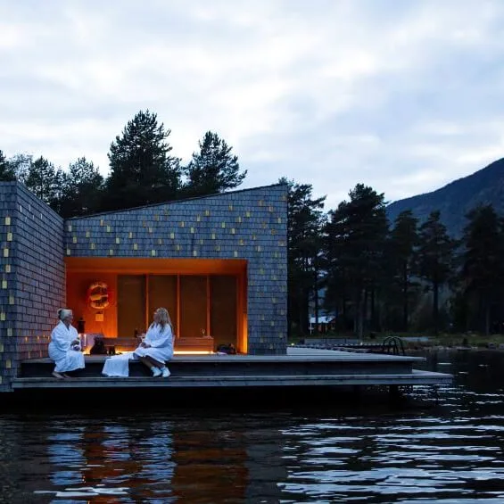 English: A modern building with angular architecture floats on water. Two people sit outside wrapped in white robes. It's surrounded by trees and mountains under a cloudy sky.