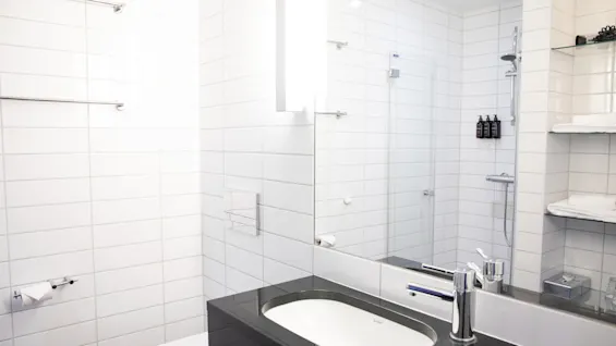 Et moderne badeværelse med en sort bordplade og håndvask, hvide flisevægge og stærke loftlys. Ved siden af er der glasskabe, en brusekabine og toiletartikler.