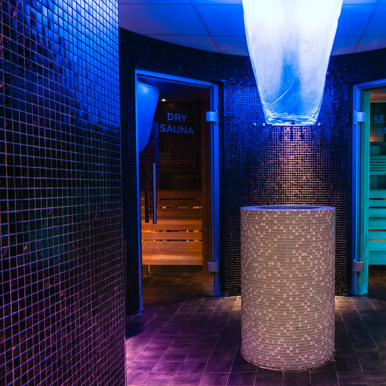 Cirkulär mosaikpelare står under blå belysning; skyltar på glasdörrar lyder "DRY SAUNA" och "SEASON SAUNA," vilket indikerar intilliggande basturum i en kaklad spamiljö. 