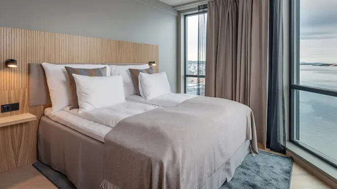 A neatly made bed with white linens and decorative pillows sits against a wooden headboard. It faces a large window with beige curtains, overlooking a scenic view.