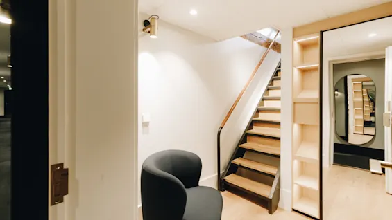 Modern hall med en trätrappa, en mörk fåtölj och inbyggda hyllor.