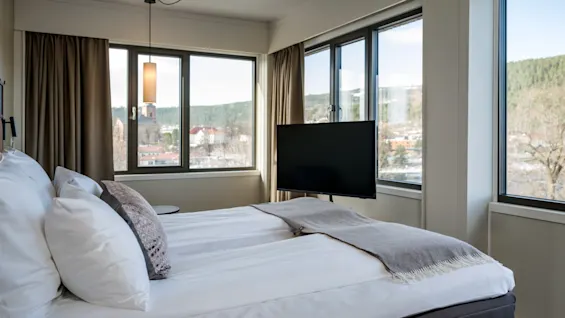 A cozy bedroom features a neatly made bed with white linens. A TV is mounted in front. Large windows provide a view of trees and buildings outside.