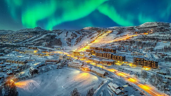 Snow-covered town illuminated by streetlights at night, with the Northern Lights dancing vibrantly in the snowy, mountainous background.