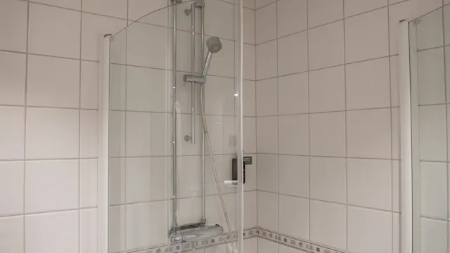 En glasinkapslad dusch står i ett badrum med vita kaklade väggar och kakel med mönstrade bårder, med ett väggmonterat justerbart duschhuvud och metallbeslag.