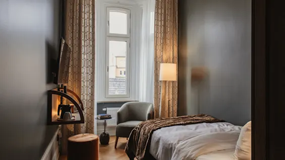 Home Hotel Baltzar - Compact Single. A cozy hotel room with a single bed, armchair, and warm lighting, perfect for relaxation.