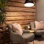 To grå lænestole med puder står ved siden af et lille rundt bord under en hængende lampe, mod en rustik trævæg med nærliggende potteplanter.