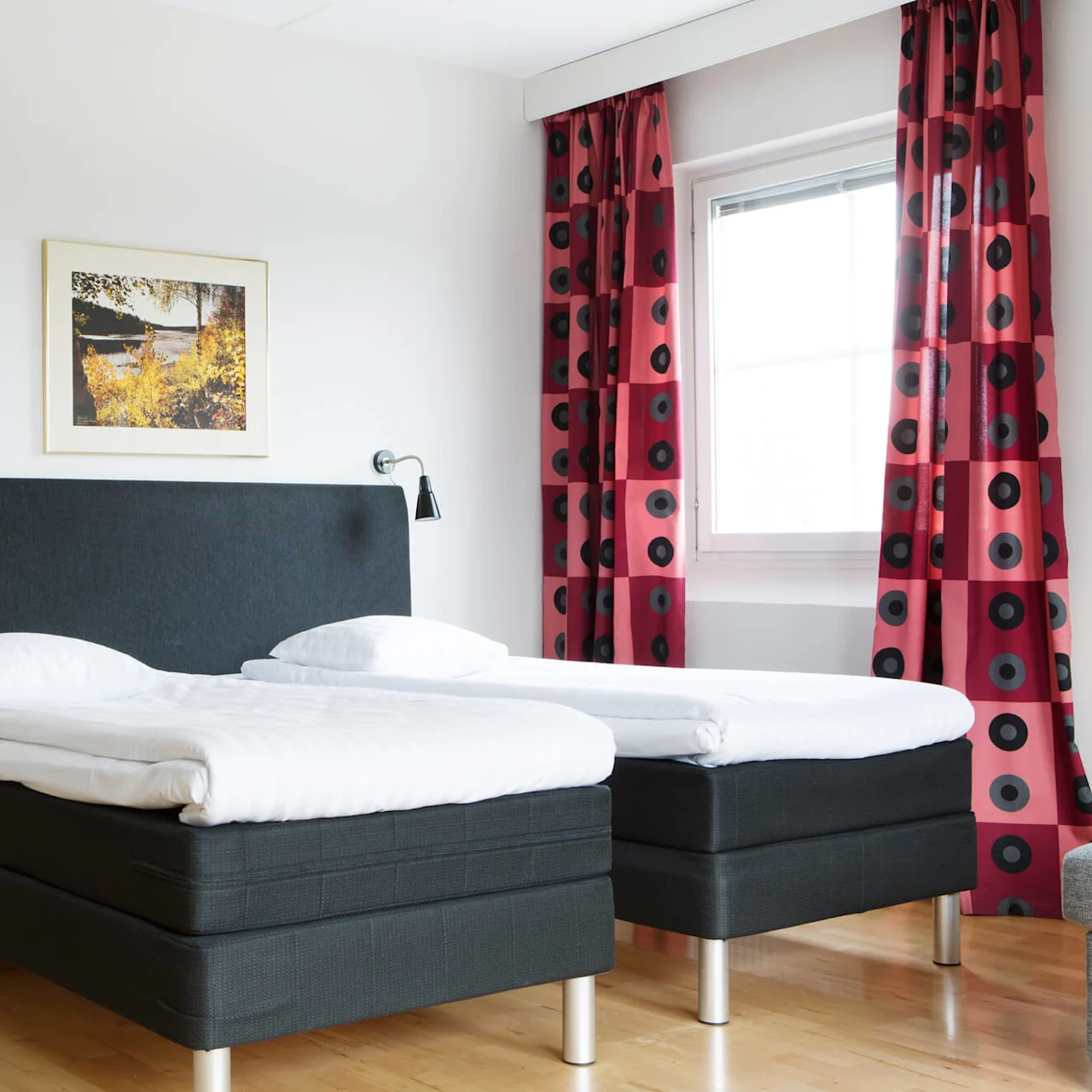 Twin beds are neatly made with white linens in a bright room. A painting hangs above the headboard, and red patterned curtains flank a window, highlighting the wooden floor.