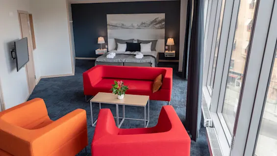 Ett rymligt hotellrum innehåller en stor säng mot en mörk vägg, omgiven av röda och orange sittmöbler. En växt står på ett soffbord, med fönster från golv till tak med utsikt över en stadsbild.