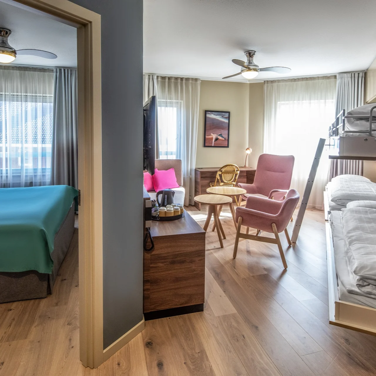 A room with a double bed and teal bedding connects to a living area through an open doorway. The living area has a pink chair, bunk beds, and wooden flooring.