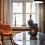Stearinlys i perforeret holder lyser på glasbord; omgivet af orange fløjlsstole i et hyggeligt, varmt belyst rum med gardiner foran vinduet.