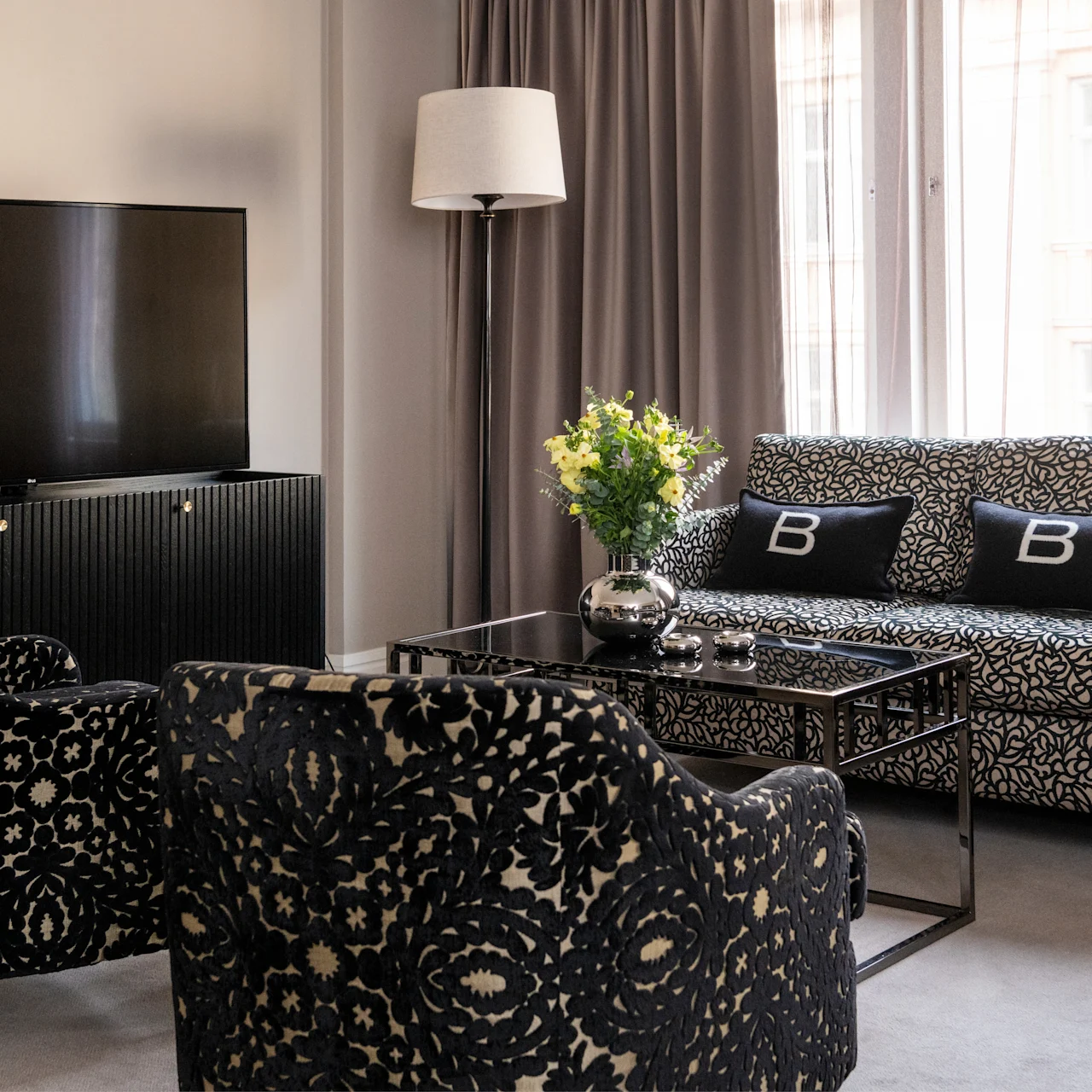 TV på et skab, med udsigt til mønstrede stole og en sofa med "B"-puder. Blomster på glasbord; vinduer med gardiner oplyser rummet.