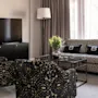 TV på et skab, med udsigt til mønstrede stole og en sofa med "B"-puder. Blomster på glasbord; vinduer med gardiner oplyser rummet.