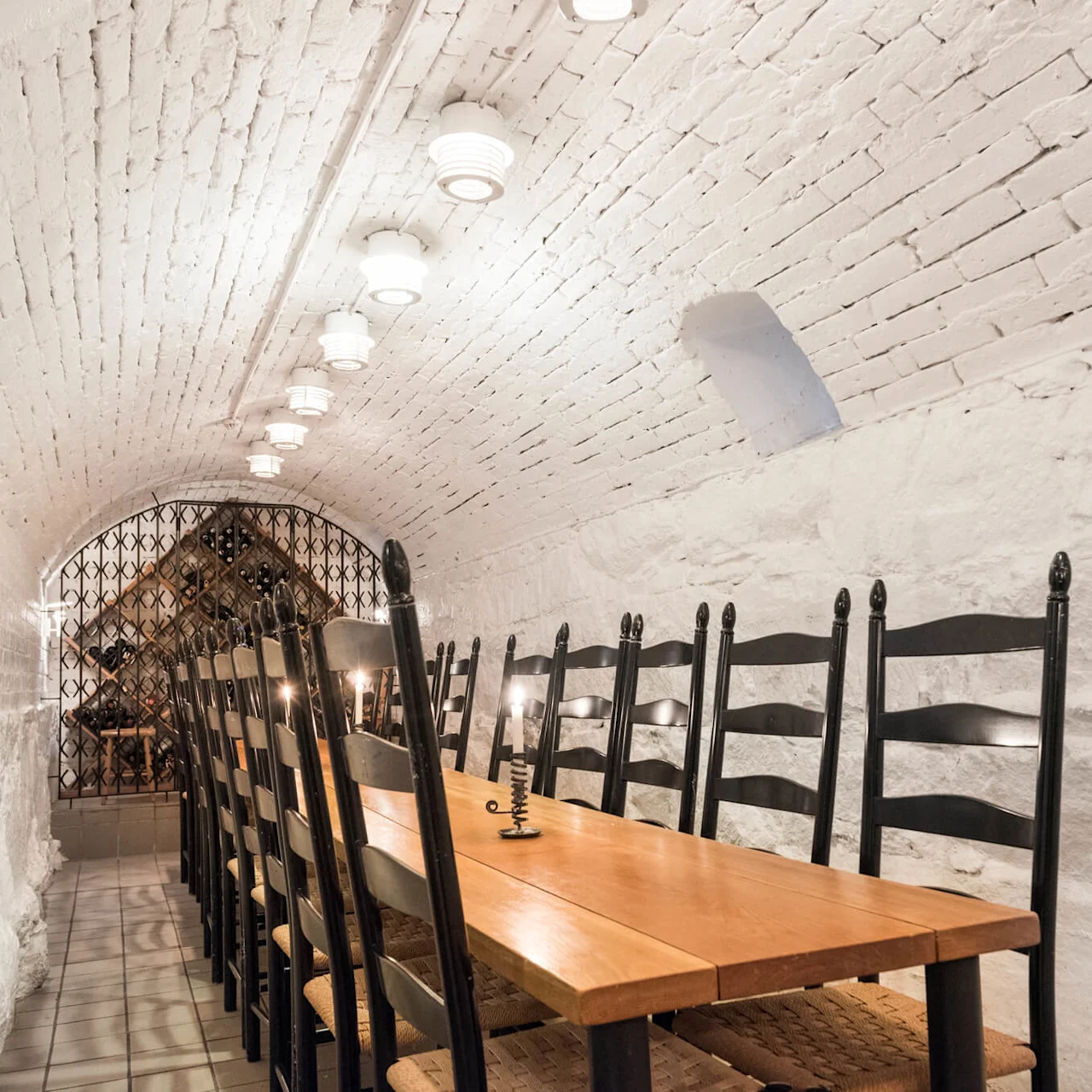 Wooden table with chairs in a white brick-vaulted cellar with wine bottles and lighting.