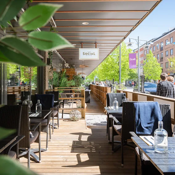En solig uteservering med dukade bord, en skylt där det står Social, och människor som promenerar förbi på gatan.