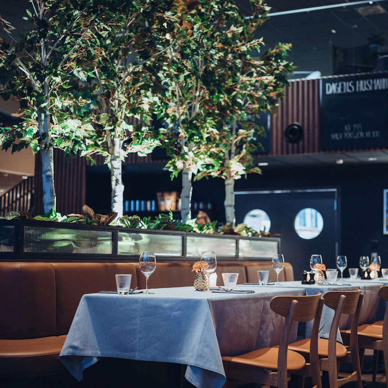 A table set with glasses and cutlery stands in a dimly lit restaurant, surrounded by wooden chairs and tall indoor plants. A menu board reads: "DAGENS HUSMAN" and "DESSERTER".