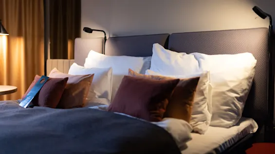 A neatly made bed features several pillows and a gray blanket. Bedside lamps illuminate the setup, and a "Please Do Not Disturb" sign hangs on a pillow. Brown curtains add warmth to the room.