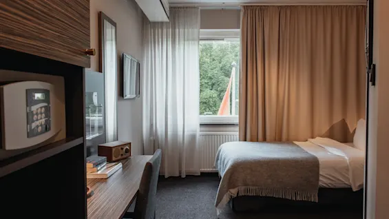 Hotellihuone, jossa on sänky, työpöytä, tallelokero ja ikkuna, jossa on ohuet ja pimennysverhot, tarjoten näkymän vehreyteen.
