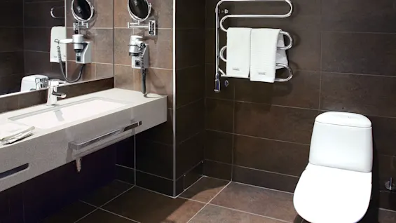 Toilet positioned beside wall-mounted radiator with towels; adjacent mirror, sink, and hairdryer; sleek, modern dark-tiled bathroom setting.