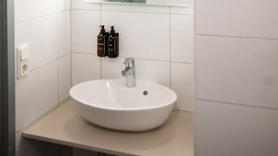 Handfat med en modern kran är placerat under en väggmonterad spegel i ett kaklat badrum. Två tvålpumpar finns ovanför. Text på spegeln lyder: "TRAPPED IN THE CLOSET."