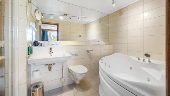 A modern bathroom with a sink, toilet, bidet, and jacuzzi tub, tiled in light beige; a window provides natural light.