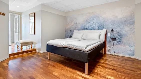 A bed with white bedding is centered against an abstract blue wall mural in a bright bedroom with wood flooring.
