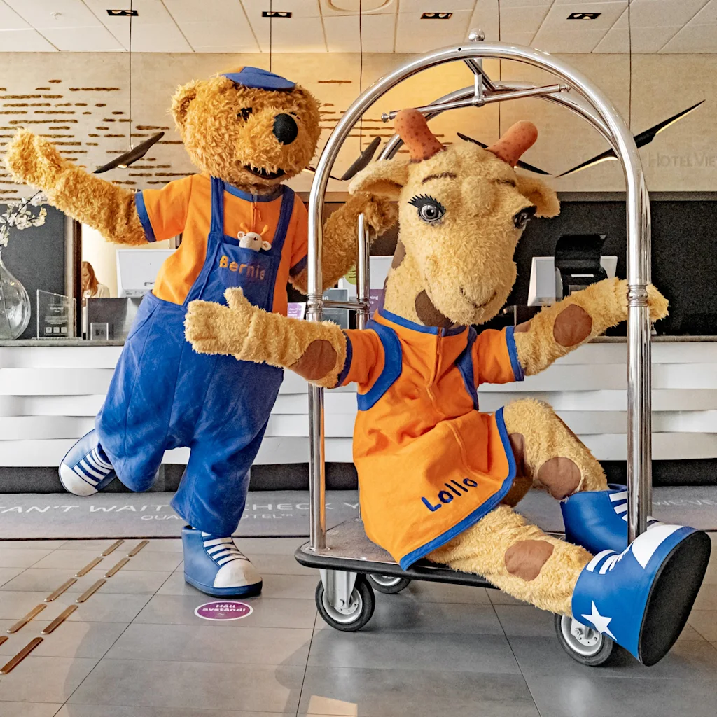 Två utklädda maskotar, en björn vid namn Bernie och en get vid namn Lollo, poserar lekfullt i en hotellobby. Bernie står, medan Lollo sitter på en bagagevagn.