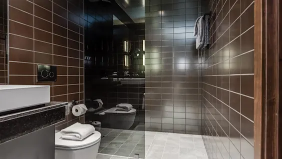 Et moderne badeværelse med brune flisevægge har et toilet med et sammenfoldet håndklæde, en sort toiletpapirholder og et væghængt håndklæde. En glasdør adskiller bruseren. 