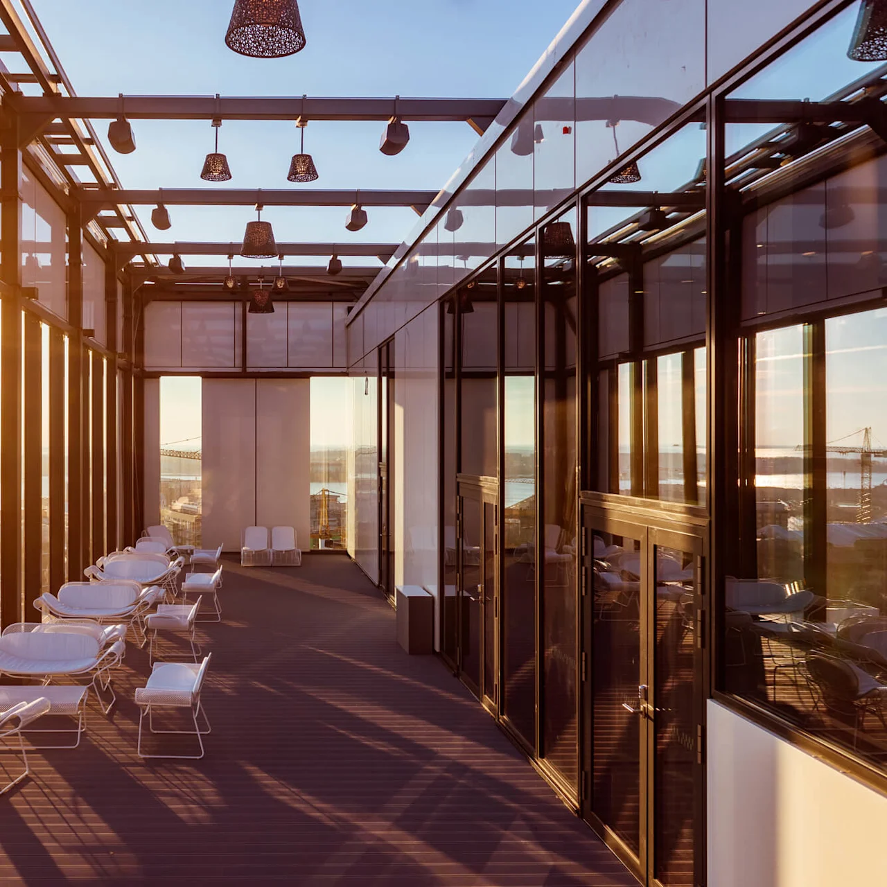Terrasse med hvide stole bader i gyldent solnedgangslys. Omgivet af glasvægge og loftlamper er bylandskabet og kraner synlige i det fjerne.