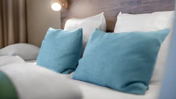 A comfortable bed features two blue pillows resting on white cushions, placed in a well-lit room with a wood headboard and soft ambient lighting from a wall lamp.