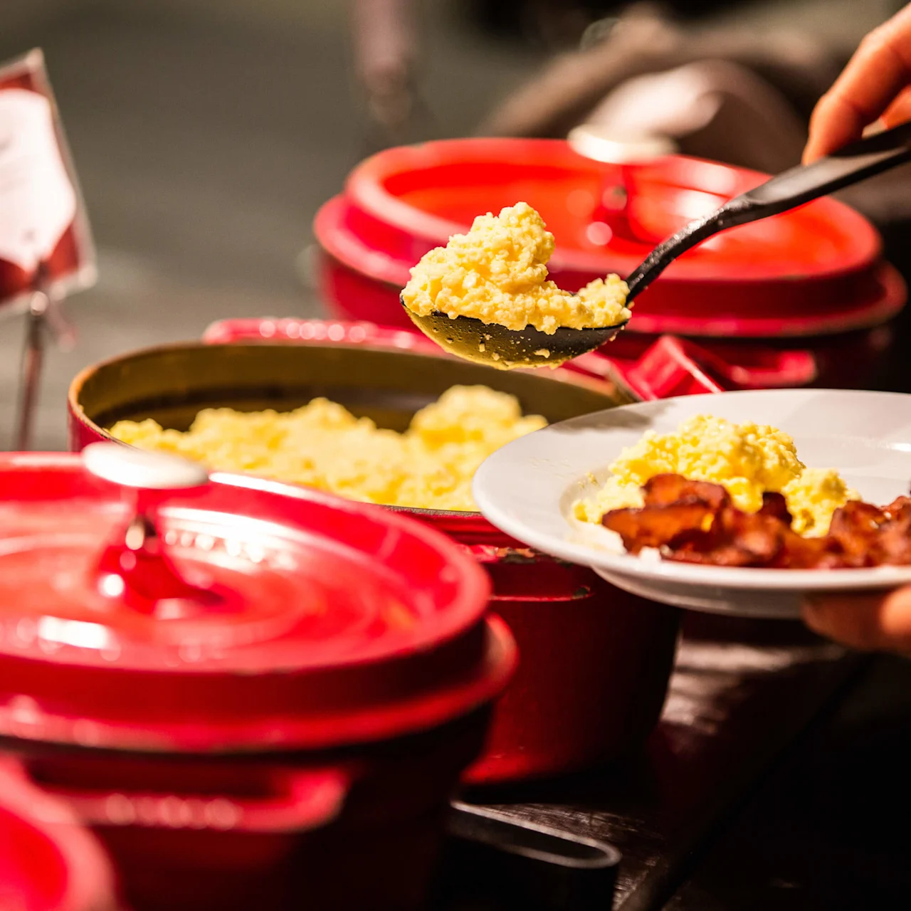 A person serves scrambled eggs onto a plate with bacon in a buffet setting at Copperhill Mountain Lodge hotel in Swedish Are..