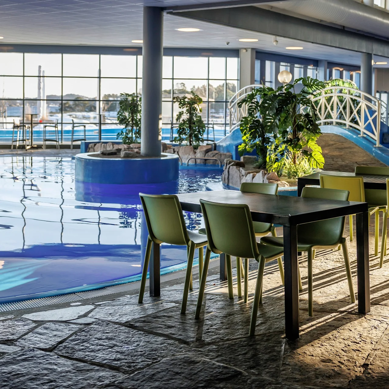 A pool with clear water reflects sunlight indoors. Nearby, tables with green chairs stand on a stone floor. Green plants and a white decorative bridge enhance the ambiance.