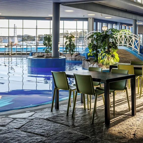 Et basseng med klart vann reflekterer sollys innendørs. I nærheten står bord med grønne stoler på et steingulv. Grønne planter og en hvit dekorativ bro forbedrer stemningen.