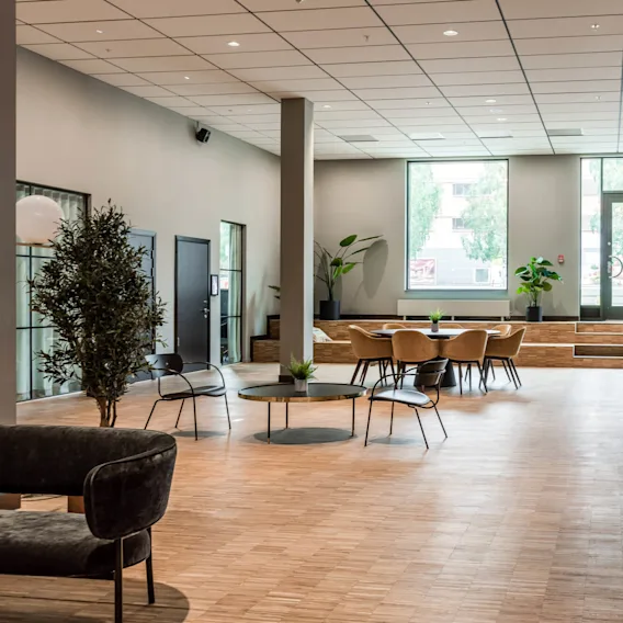 A spacious modern lobby features a seating area with round tables and chairs, surrounded by potted plants. A counter with coffee machines lines the right wall.