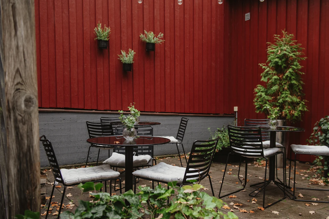 A cozy outdoor seating area with black tables and chairs against a red wooden wall, adorned with plants and fallen leaves. Home Hotel Kung Oscar.