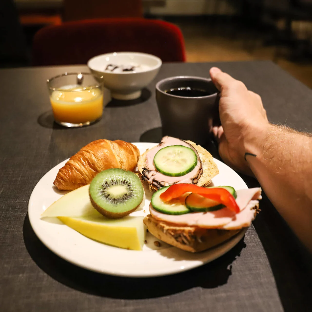 A breakfast plate featuring a croissant, slices of cheese, kiwi, and sandwiches topped with cold cuts, cucumber, and bell pepper. A hand is holding a cup of black coffee.