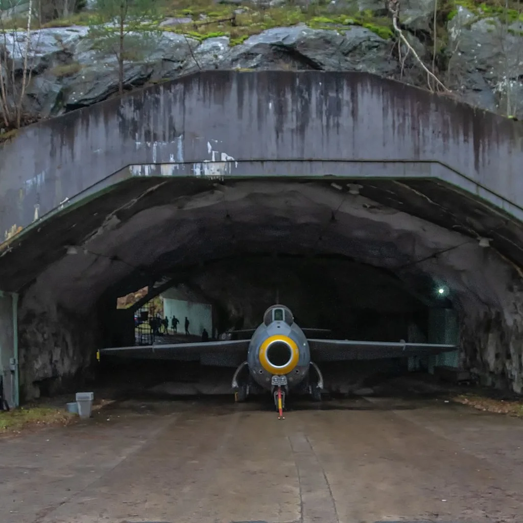 Et veteranjetfly er placeret ved indgangen til en betonhangar bygget ind i en klippefyldt, mosbegroet bakke.
