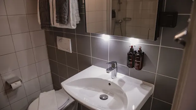 Handfat med blandare är belyst under en spegel i ett kaklat badrum. Väggmonterade tvåldispenser finns i närheten och handdukar är vikta på en ställning. Dusch synlig i bakgrunden.