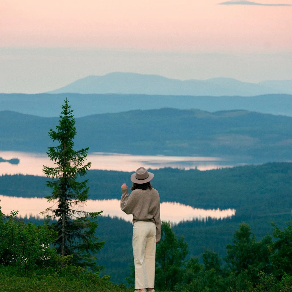 En person med hat står på en bakke og ser ud over et fredfyldt landskab med skove, en rolig sø og fjerne bjerge ved solnedgang.