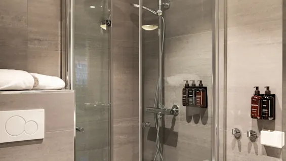 Ett modernt badrum med en duschkabin i glas, grå kaklade väggar och väggmonterade toalettartiklar. Färska vita handdukar syns.