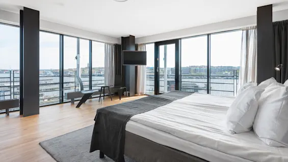 Bed with white sheets and gray blanket sits in a modern, spacious room. Large windows offer a panoramic city view, and a wall-mounted TV faces the bed.