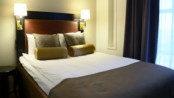 A neatly made double bed with pillows and lamps on either side, in a hotel room.