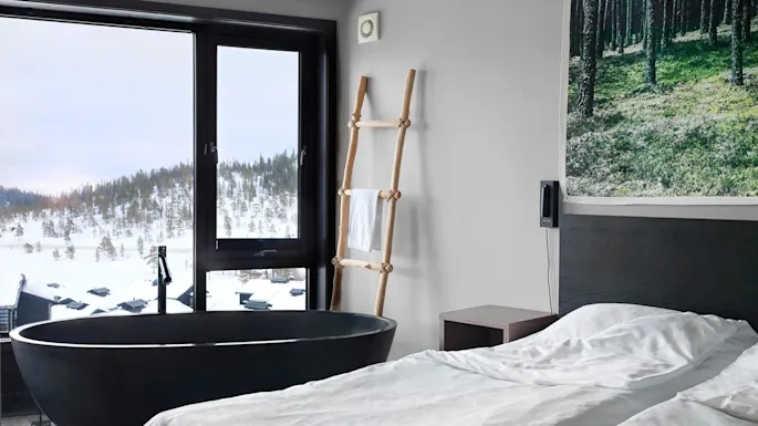 A modern bedroom features a bed, a freestanding bathtub, and a wood ladder with a towel, set against a window showcasing a snowy landscape and forested hills.