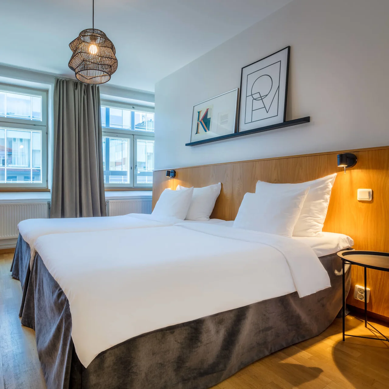A neatly-made bed in a well-lit, modern hotel room with wooden headboard, artwork on the wall, and simplistic furniture.